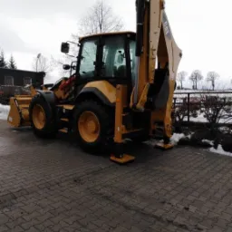 Żółta koparko-ładowarka CAT zaparkowana na brukowanej powierzchni z wysuniętymi stabilizatorami, widok z tyłu, pochmurne niebo w tle.