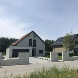 Dwa nowoczesne domy jednorodzinne z płaskimi dachami i elewacją w odcieniach szarości, ogrodzone niskim betonowym murem, z numerem 51 na jednym z budynków.