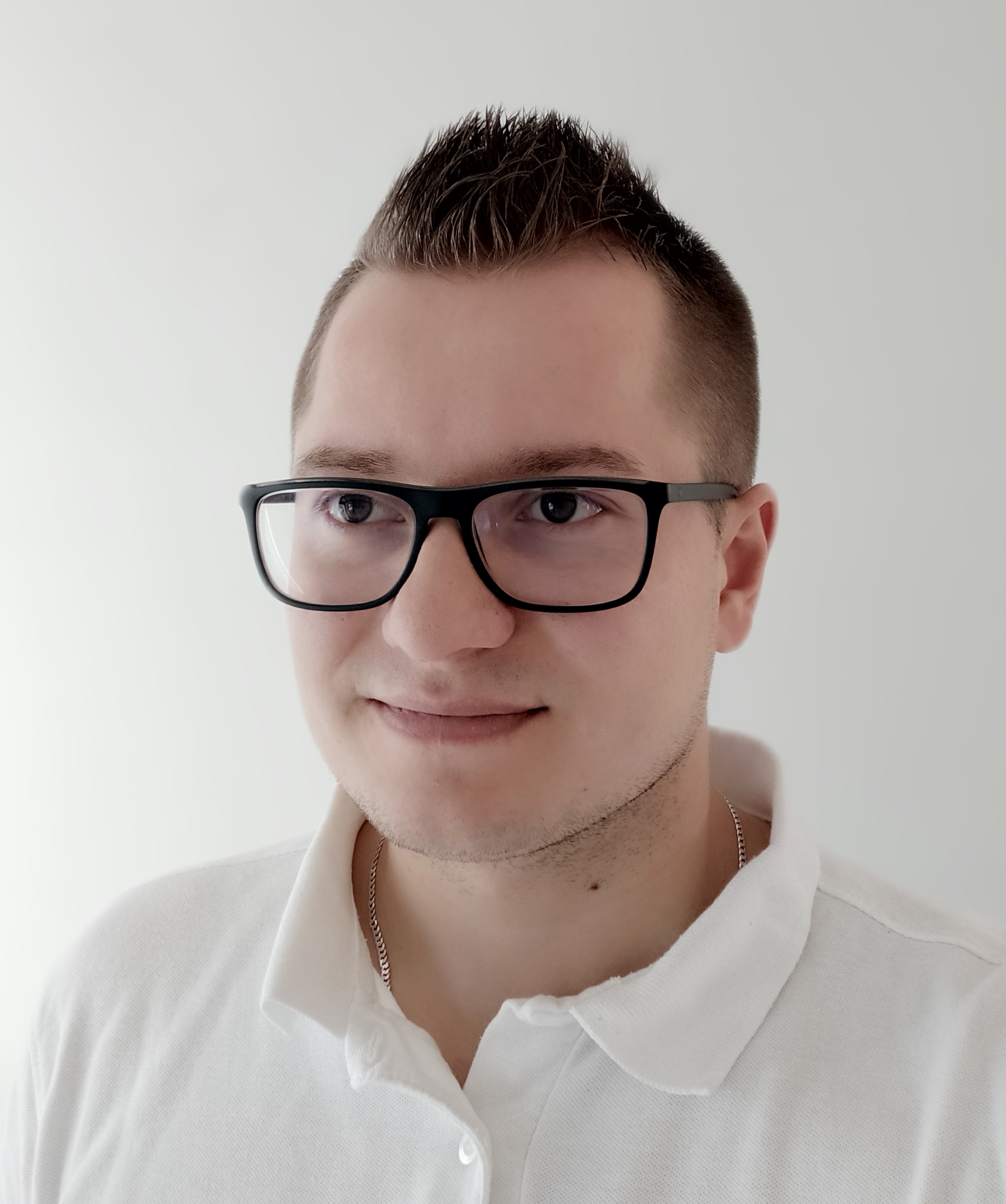 Portret mężczyzny w okularach z krótką fryzurą i srebrnym łańcuszkiem, ubranego w białą koszulkę polo na tle jasnej ściany.