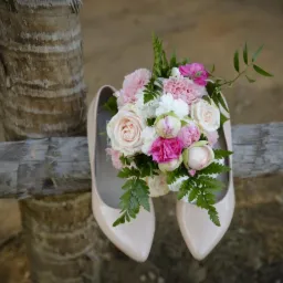 Beżowe szpilki z bukietem różowych i białych kwiatów leżące na drewnianej belce przy pniu drzewa.