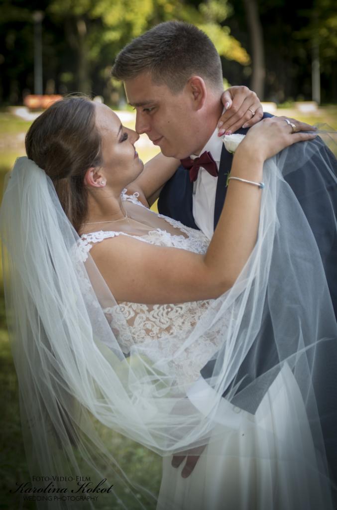 Młoda para w objęciach, zbliżająca się do pocałunku w plenerze; panna młoda w koronkowej sukni i welonie, pan młody w granatowym garniturze z bordową muchą.