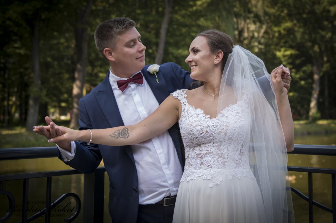 Uśmiechnięta para młoda w objęciach na tle zieleni parkowej, panna młoda w koronkowej sukni z welonem, pan młody w granatowym garniturze z bordową muszką, widoczny tatuaż na ramieniu panny młodej.