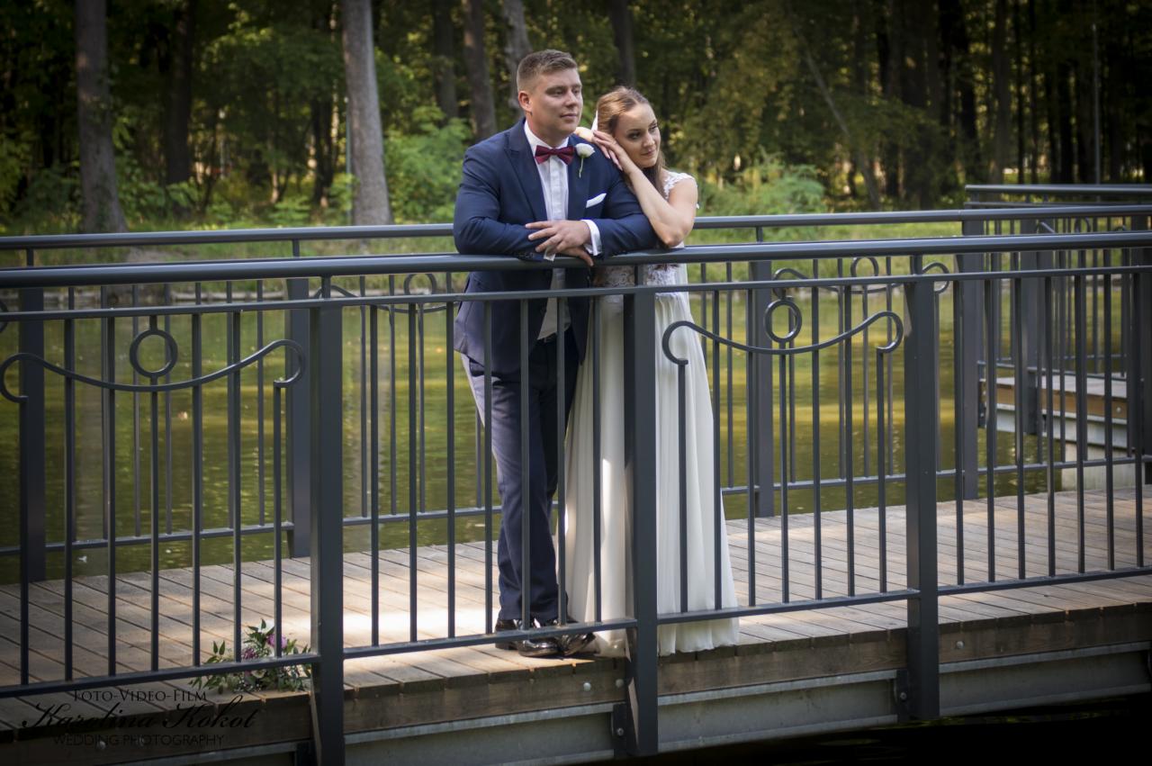 Para młoda w eleganckich strojach pozuje na moście z metalową balustradą na tle zieleni i wody.