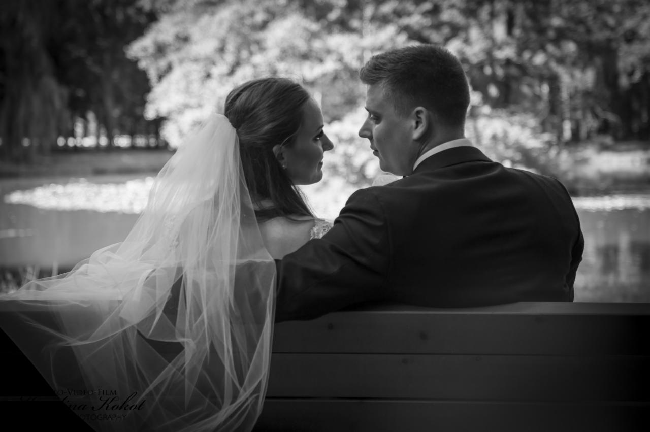 Czarno-białe ujęcie pary młodej siedzącej na ławce, z profilu, spoglądającej na siebie z czułością, z jeziorem i drzewami w tle.