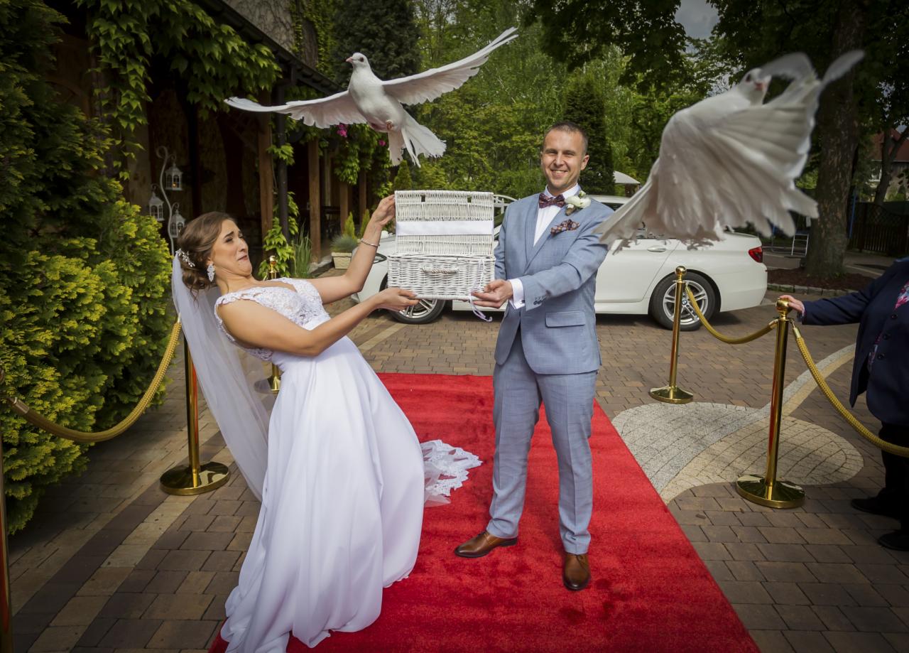 Para młoda na czerwonym dywanie wypuszcza białe gołębie z wiklinowego koszyka przed budynkiem z zielonymi pnączami.