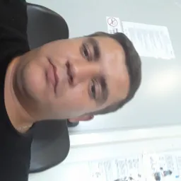 Portret mężczyzny w czarnej koszulce na tle białej ściany z plakatem informacyjnym i znakiem zakazu palenia. Ujęcie z bliska, lekko z dołu.