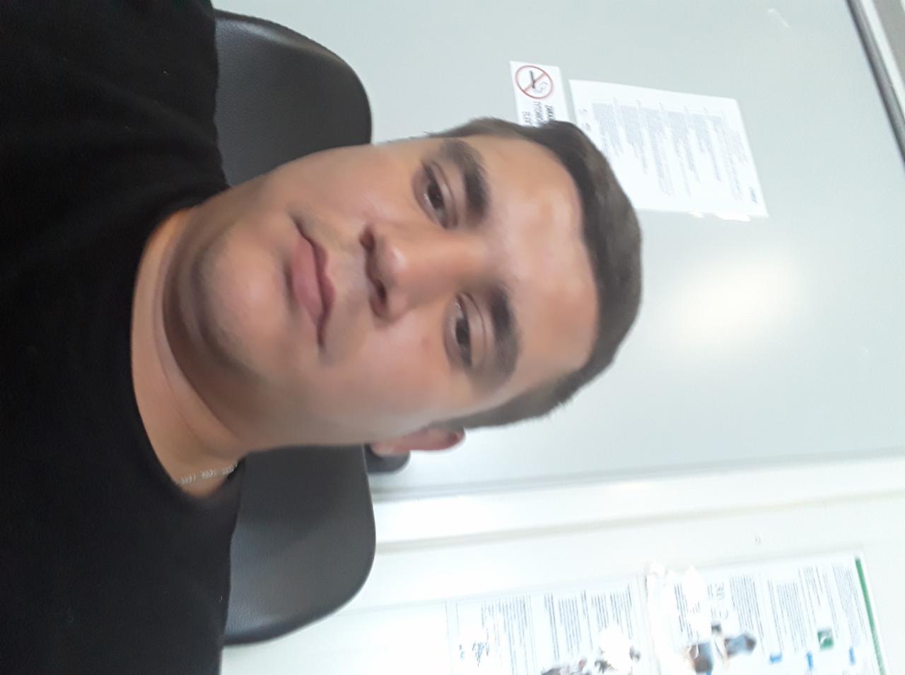 Portret mężczyzny w czarnej koszulce na tle białej ściany z plakatem informacyjnym i znakiem zakazu palenia. Ujęcie z bliska, lekko z dołu.