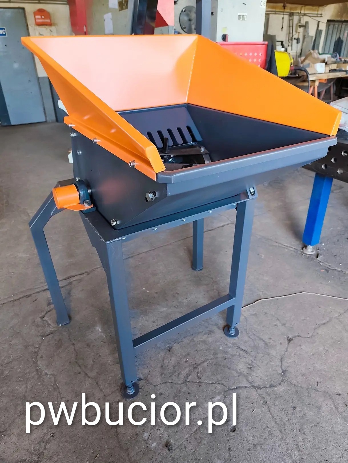 Szara sieczkarnia z pomarańczowym podajnikiem, spawana konstrukcja na czterech nogach, widoczne śruby i elementy mechaniczne. Nowoczesny design w warsztatowym otoczeniu.