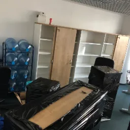 Biuro w trakcie pakowania: meble i sprzęt zabezpieczone czarną folią stretch, w tle regał z półkami i butle z wodą.