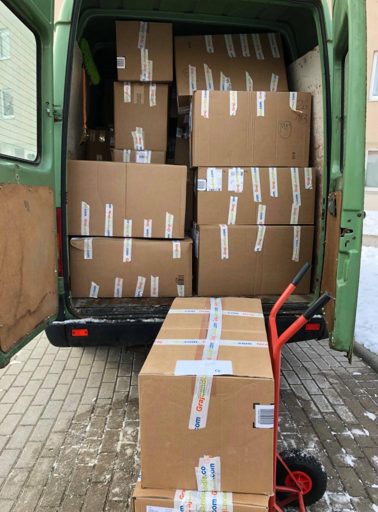 Wnętrze zielonego busa załadowane kartonowymi pudłami, część z nich zabezpieczona taśmą z napisem, na zewnątrz widać wózek transportowy z kolejnymi kartonami, zimowy krajobraz w tle.