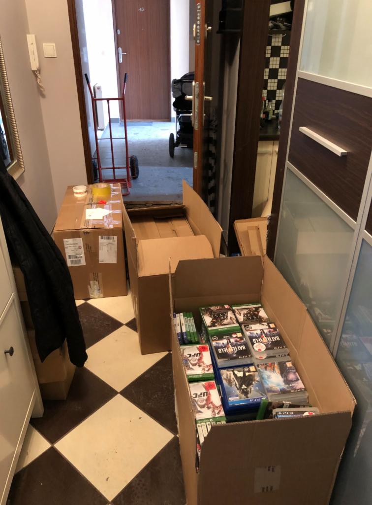Korytarz z kartonami pełnymi gier wideo, w tle wózek transportowy, przygotowania do przeprowadzki.