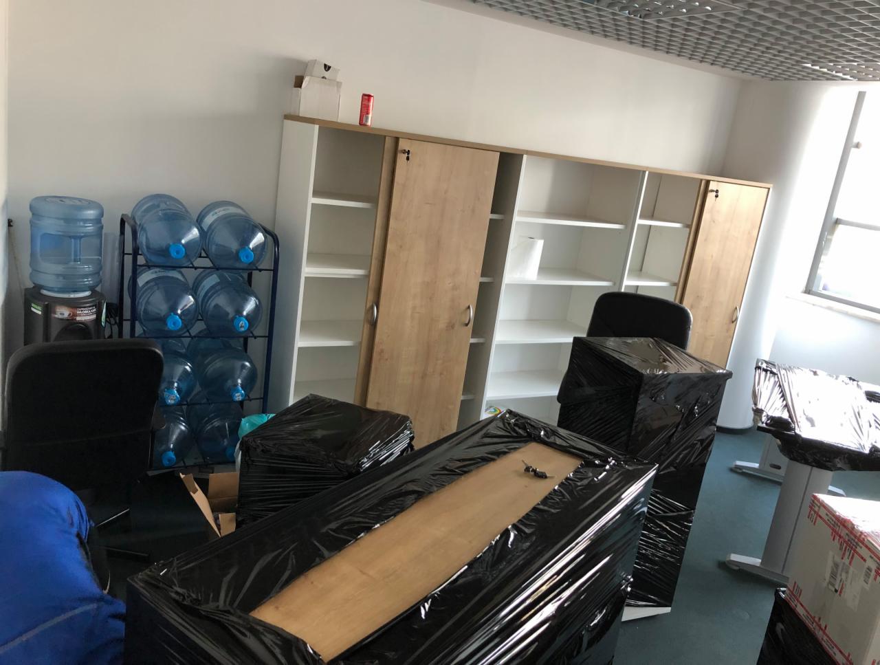Biuro w trakcie pakowania: meble i sprzęt zabezpieczone czarną folią stretch, w tle regał z półkami i butle z wodą.