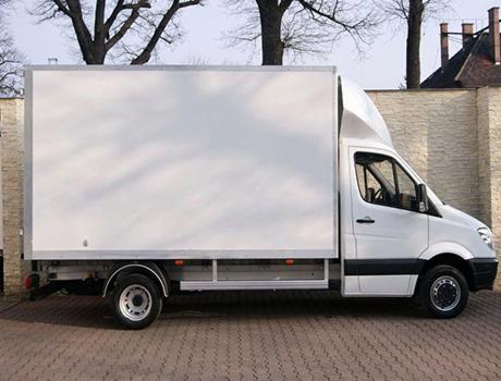 Biały samochód dostawczy typu furgon z wysoką kabiną, zaparkowany na brukowanej powierzchni, z widocznymi drzewami bez liści w tle i fragmentem ceglanego budynku.