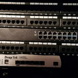 Panel krosowniczy z przełącznikiem sieciowym Ubiquiti EdgeSwitch i routerem DrayTek Vigor zamontowane w szafie rack, widoczne porty RJ45 i oznaczenia kategorii Cat.6.