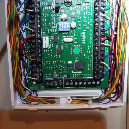 Otwarta skrzynka centrali alarmowej Texecom z podłączonymi kolorowymi przewodami, widoczne złącza, układy scalone i przełączniki DIP.