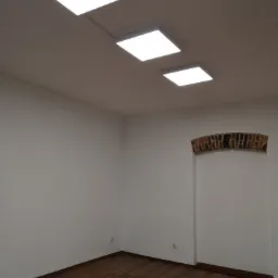 Wnętrze pomalowanego pomieszczenia z trzema kwadratowymi lampami LED na suficie i drewnianą podłogą, widoczne gniazdka elektryczne na ścianie oraz fragment łuku z cegły nad otworem drzwiowym.