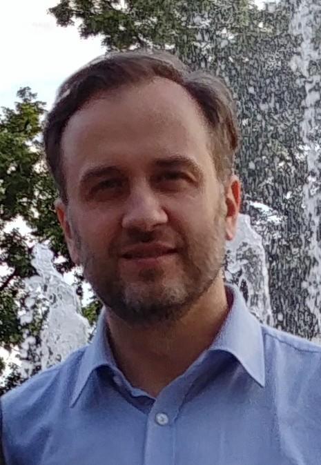 Portret mężczyzny w błękitnej koszuli na tle fontanny i zieleni, z delikatnym uśmiechem i lekkim zarostem. Ujęcie od przodu, skupione na twarzy.