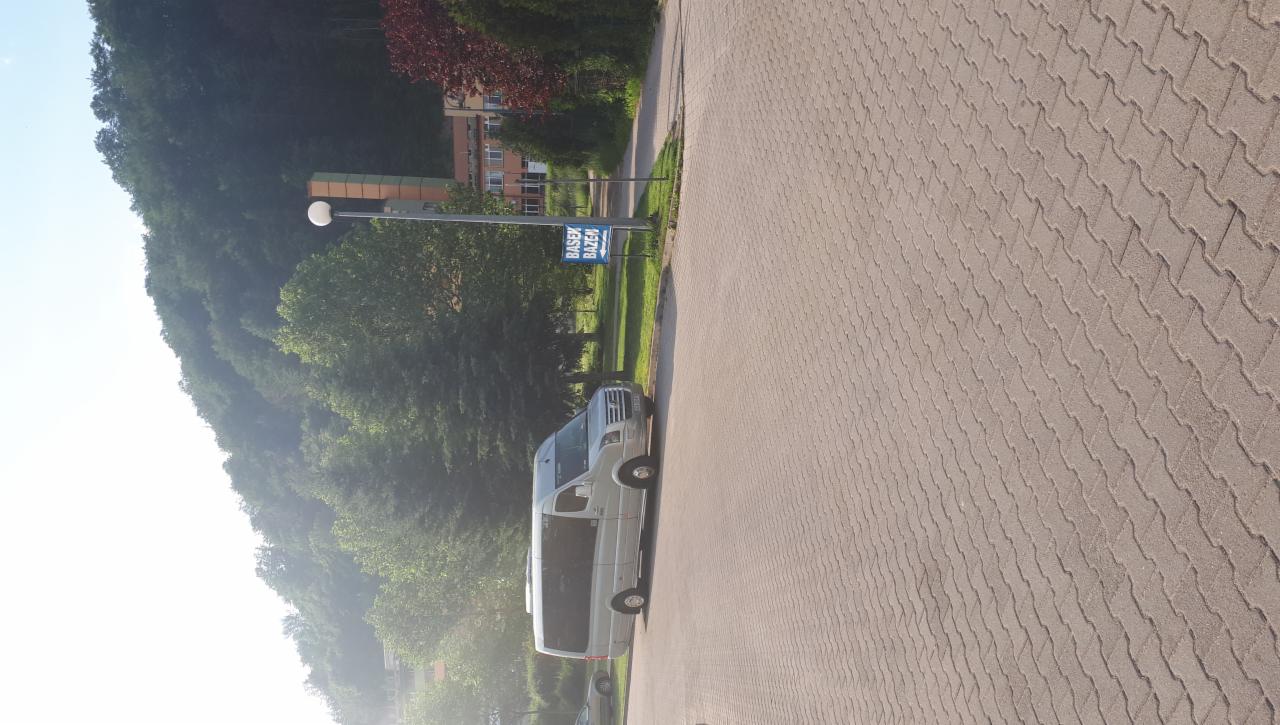 Biały bus zaparkowany na brukowanym parkingu obok zieleni i drogowskazu 'Basen Bazen'.