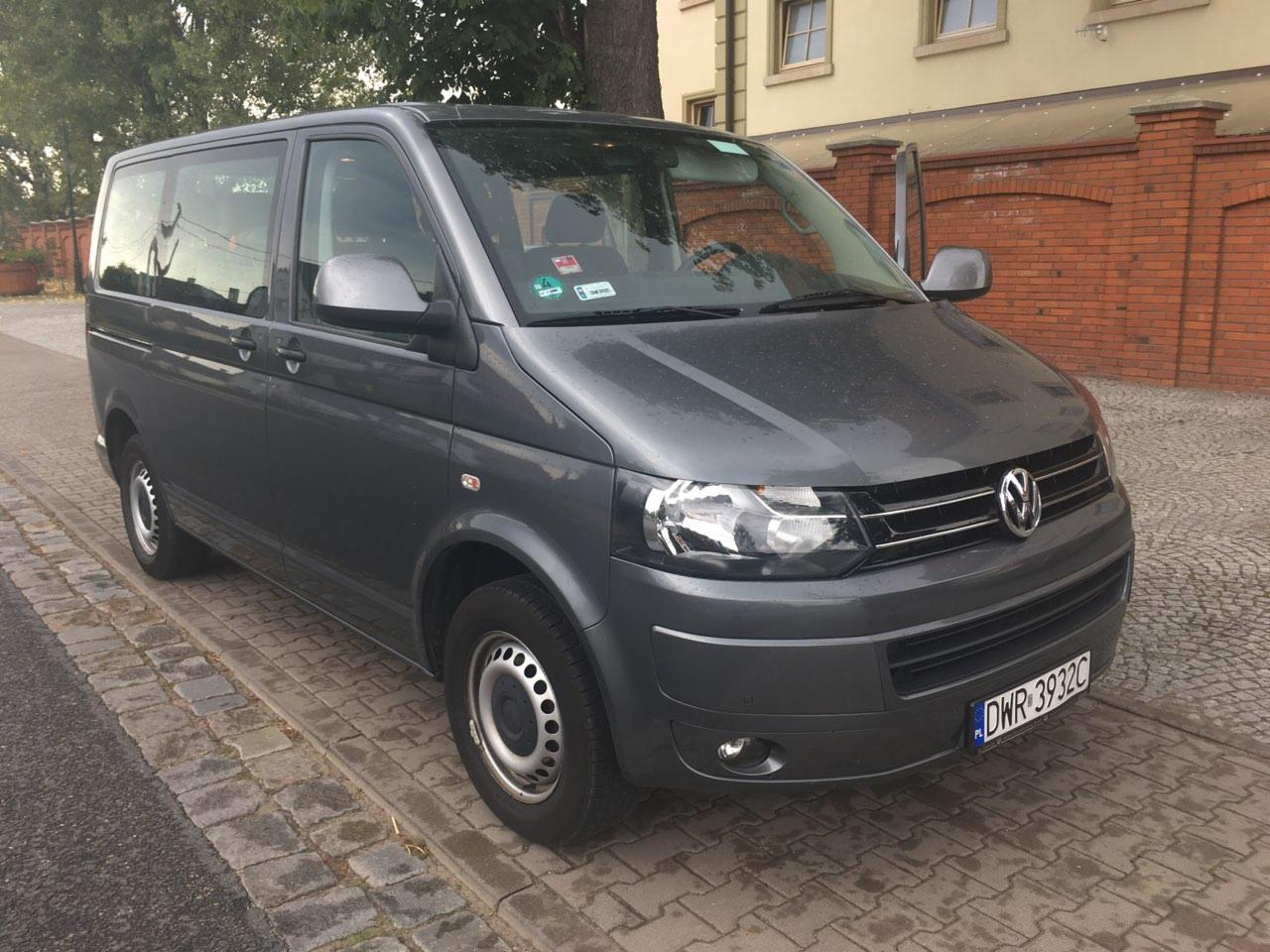 Szary bus Volkswagen Transporter zaparkowany na brukowanej ulicy przed budynkiem z cegły.