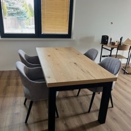 Meble Damian Góralski - Drewniany stół z czarnymi nogami i szarymi krzesłami w nowoczesnym wnętrzu. Widok na okno z żaluzjami i fragment wyposażenia. Parkiet na podłodze.