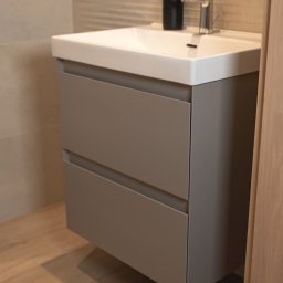 Meble Damian Góralski - Szara szafka pod umywalkę z dwiema szufladami, biała umywalka, chromowana bateria i czarny dozownik na tle beżowej ściany z poziomymi paskami.