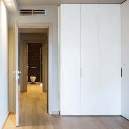 Meble Damian Góralski - Biała szafa wnękowa z minimalistycznymi uchwytami, widok z korytarza z otwartymi drzwiami do łazienki z brązową mozaiką i drewnianą podłogą.