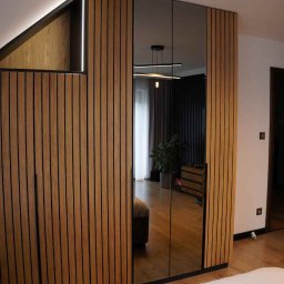 Meble Damian Góralski - Szafa wnękowa z lustrzanymi drzwiami i drewnianymi lamelami w nowoczesnej sypialni na poddaszu. Widoczne oświetlenie LED i odbicie wnętrza pokoju.