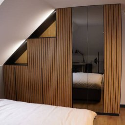 Meble Damian Góralski - Szafa wnękowa z lustrzanymi drzwiami i drewnianymi lamelami, dopasowana do skosu poddasza, widoczna z perspektywy łóżka w sypialni.