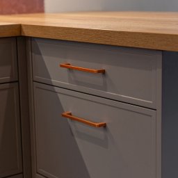Meble Damian Góralski - Szare fronty szuflad z miedzianymi uchwytami pod blatem z jasnego drewna. Minimalistyczny design, nowoczesne wykończenie.