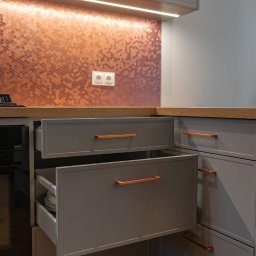 Meble Damian Góralski - Szare szafki kuchenne z otwartymi szufladami i miedzianymi uchwytami, na tle mozaiki w odcieniach miedzi i oświetlenia LED. Widoczny fragment blatu i podłogi.