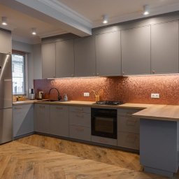 Meble Damian Góralski - Szara kuchnia na wymiar z miedzianym panelem nad blatem i drewnianym blatem. Nowoczesne oświetlenie punktowe i parkiet w jodełkę. Lodówka ze stali nierdzewnej.