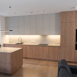 Meble Damian Góralski - Nowoczesna kuchnia z wyspą. Jasne drewno i szare szafki górne. Oświetlenie LED pod szafkami. Minimalistyczny design z czarnymi akcentami. Widok na salon.