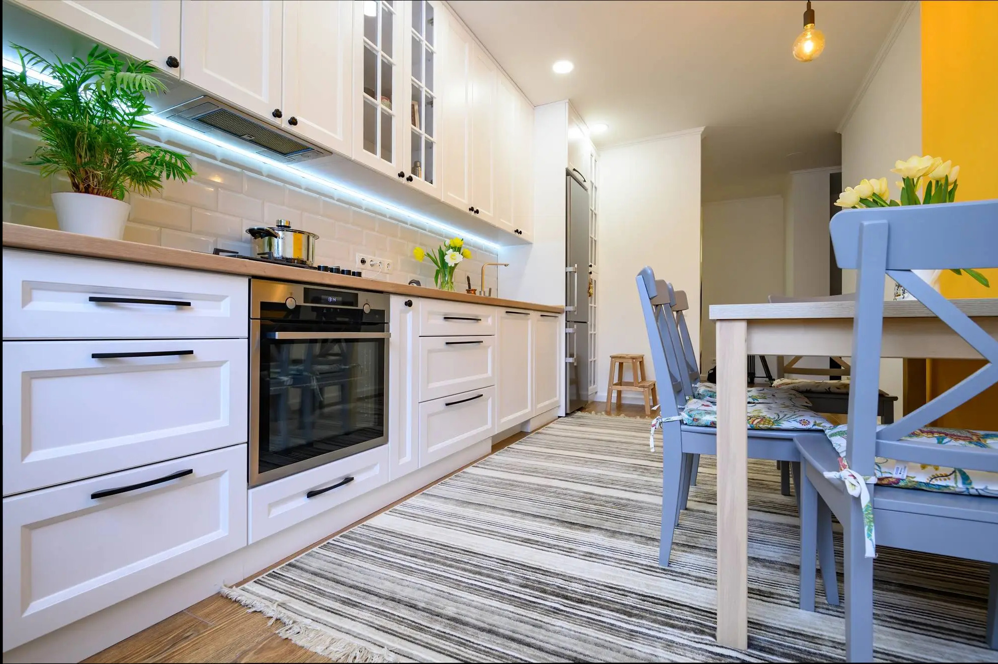 Biała kuchnia z drewnianym blatem, czarnymi uchwytami i oświetleniem LED pod szafkami. Na blacie roślina w doniczce i kwiaty w wazonie. W tle stół z krzesłami.
