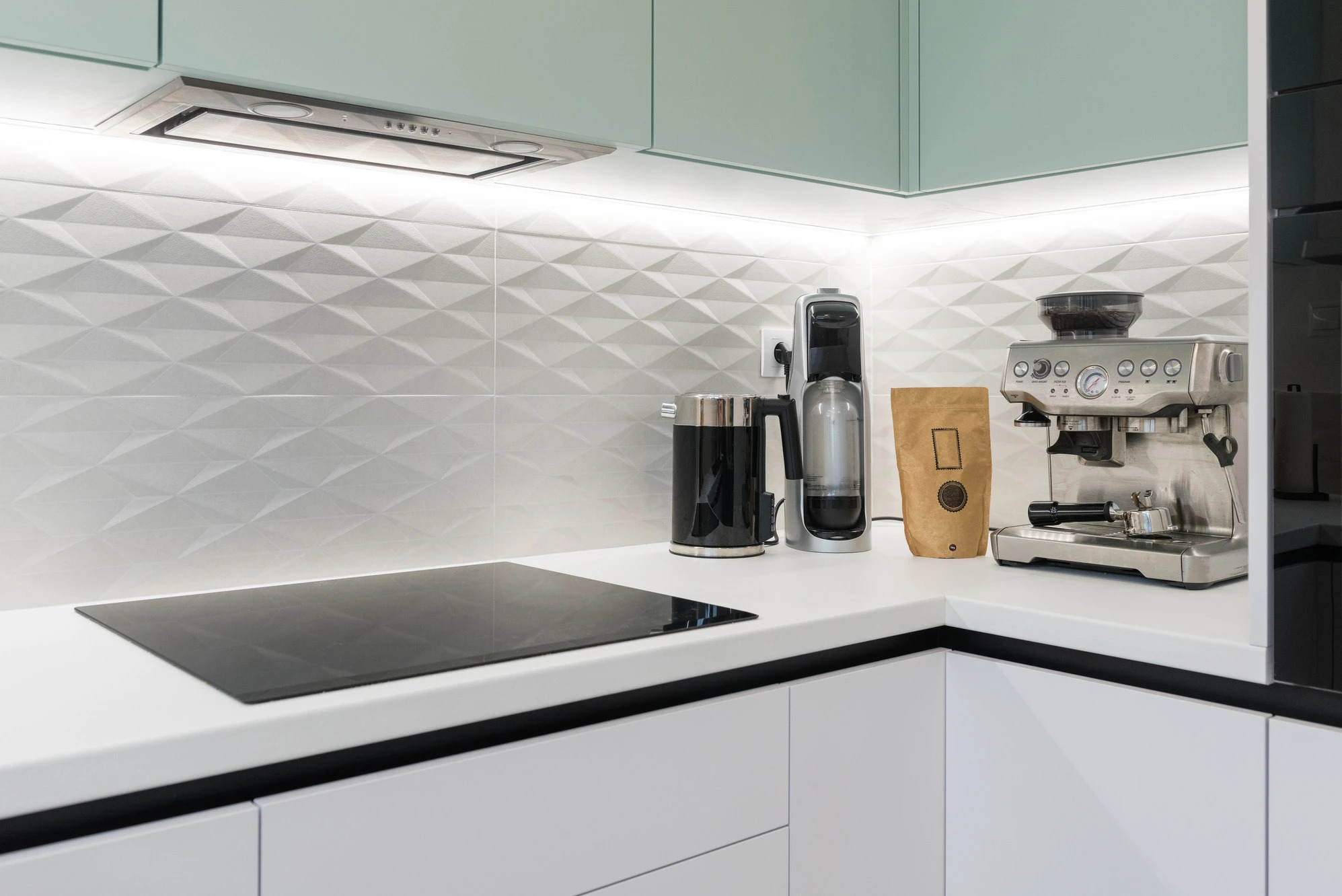 Nowoczesna kuchnia z białymi szafkami, blatem i indukcyjną płytą. Na blacie czajnik, ekspres do kawy i ozdobna ściana z trójwymiarowych płytek. Minimalistyczny design.