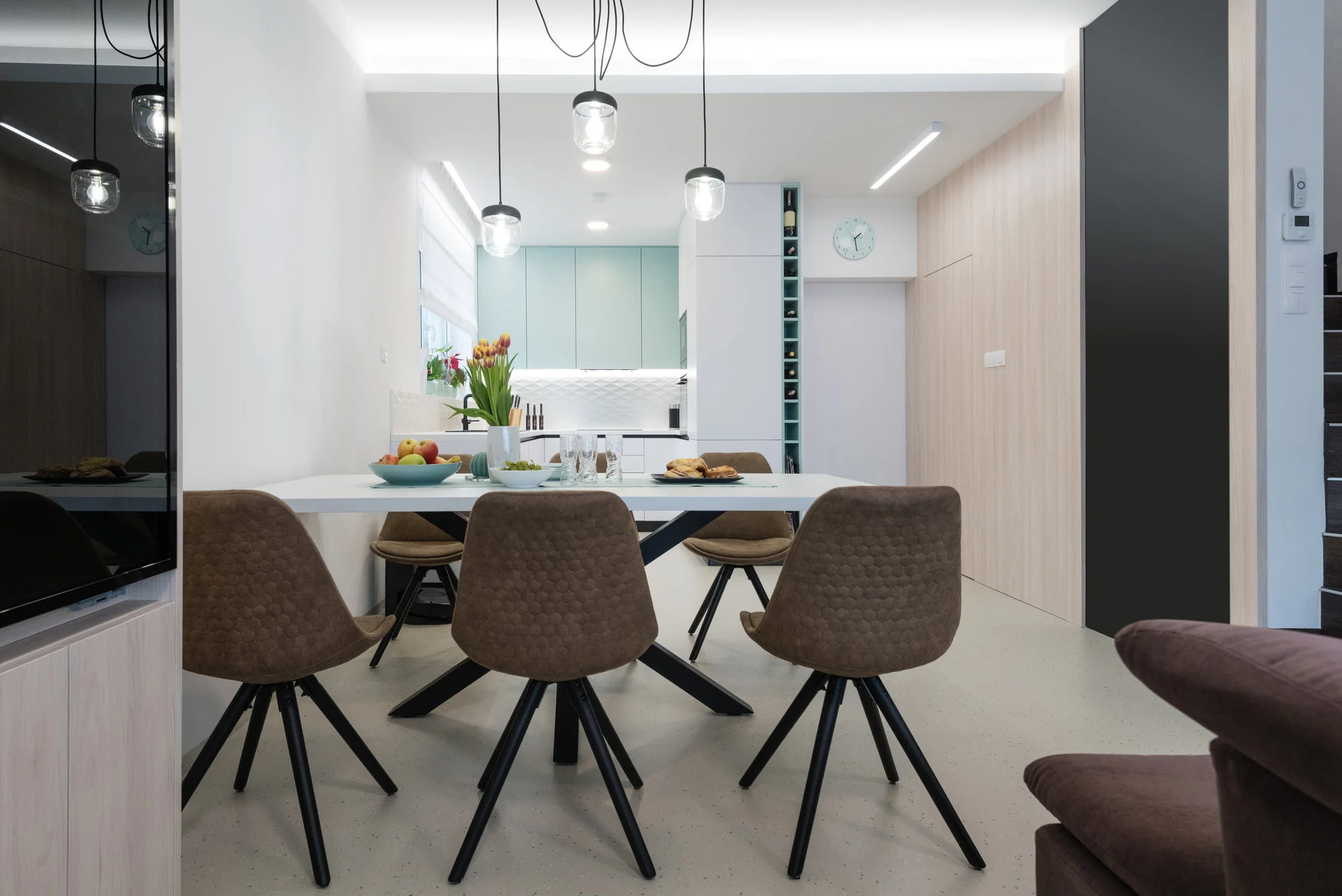 Nowoczesna kuchnia z jadalnią: biały stół z owocami i kwiatami, brązowe krzesła, jasne fronty szafek i subtelne oświetlenie. Minimalistyczny design z akcentami kolorystycznymi.
