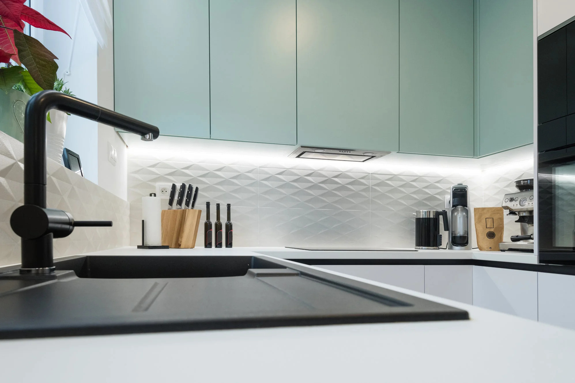 Nowoczesna kuchnia z miętową zabudową, czarnym zlewem i baterią. Białe, trójwymiarowe płytki na ścianie. Oświetlenie LED pod szafkami. Minimalistyczny design.