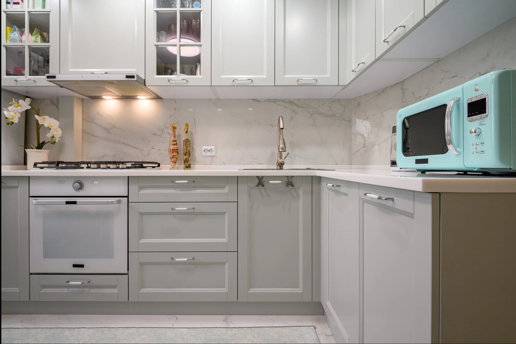Jasnoszara kuchnia na wymiar z marmurowym blatem i turkusową mikrofalówką. Górne szafki przeszklone, oświetlenie punktowe. Nowoczesny design z elementami retro.