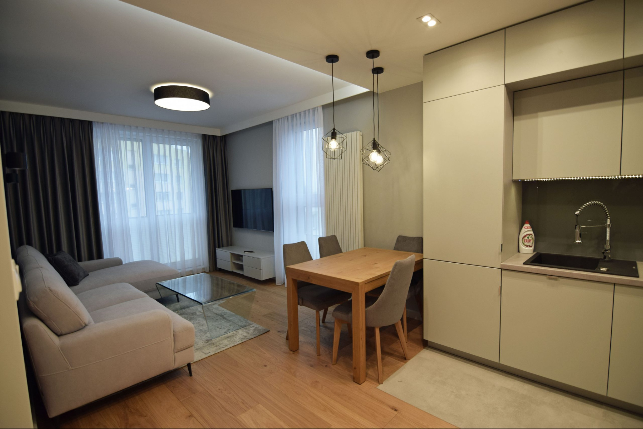 Nowoczesne wnętrze: beżowa sofa, szklany stolik, drewniany stół z krzesłami i zabudowa kuchenna w odcieniach szarości. Minimalistyczne lampy wiszące.