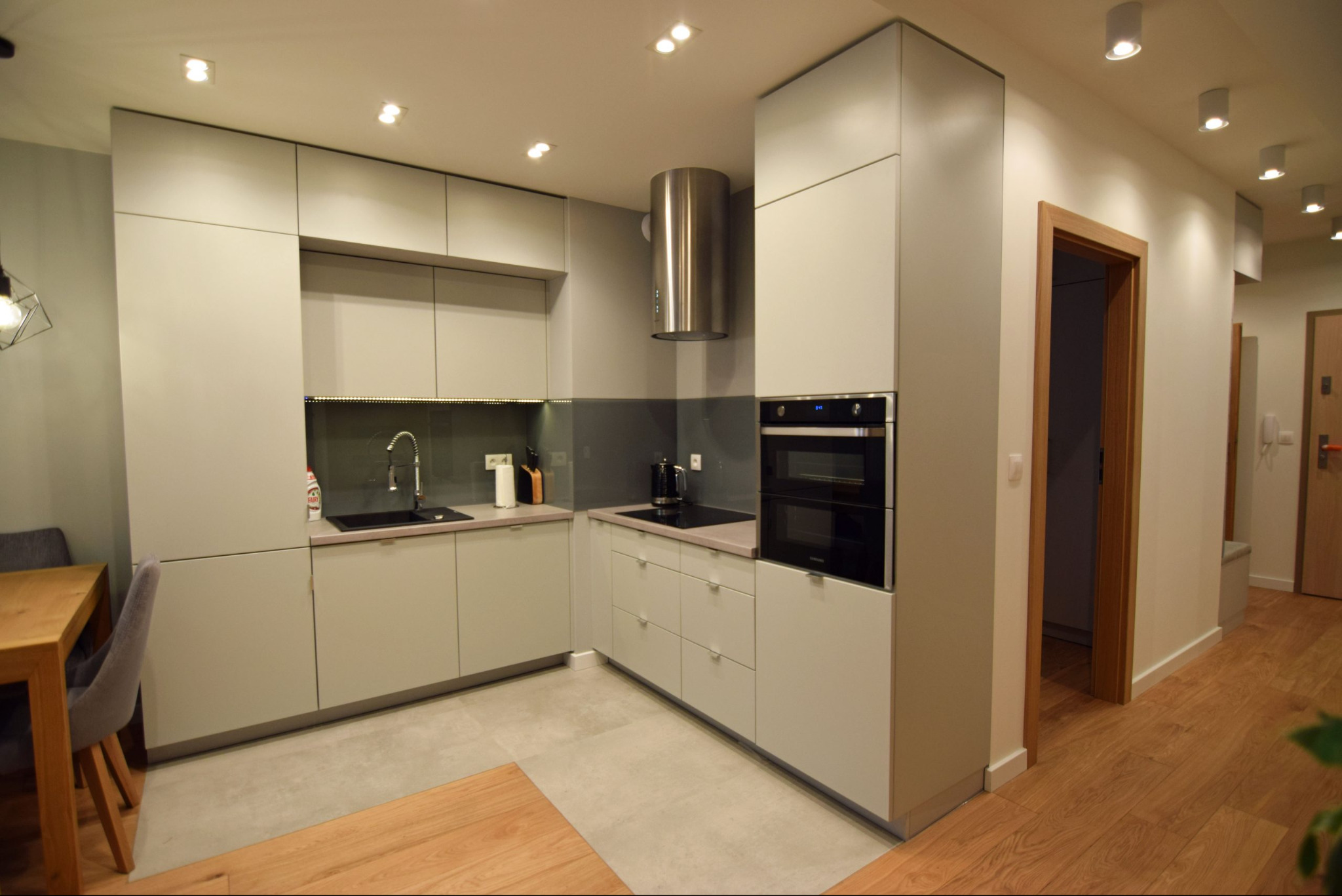 Nowoczesna kuchnia w odcieniach szarości z drewnianym stołem. Minimalistyczny design, oświetlenie punktowe i stalowy okap cylindryczny. Widok na korytarz.