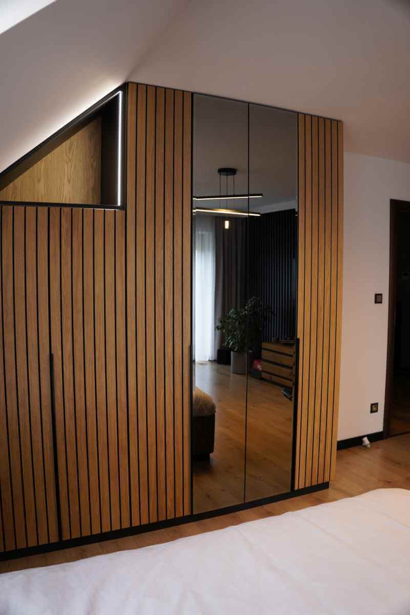 Szafa wnękowa z lustrzanymi drzwiami i drewnianymi lamelami w nowoczesnej sypialni na poddaszu. Widoczne oświetlenie LED i odbicie wnętrza pokoju.