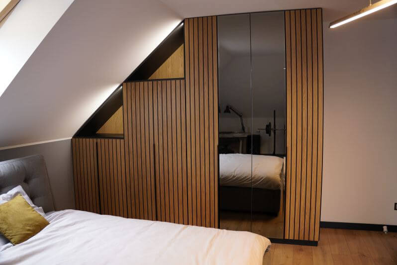Szafa wnękowa z lustrzanymi drzwiami i drewnianymi lamelami, dopasowana do skosu poddasza, widoczna z perspektywy łóżka w sypialni.