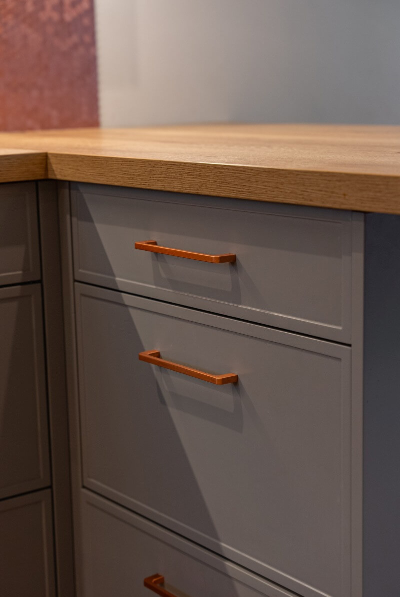 Szare fronty szuflad z miedzianymi uchwytami pod blatem z jasnego drewna. Minimalistyczny design, nowoczesne wykończenie.