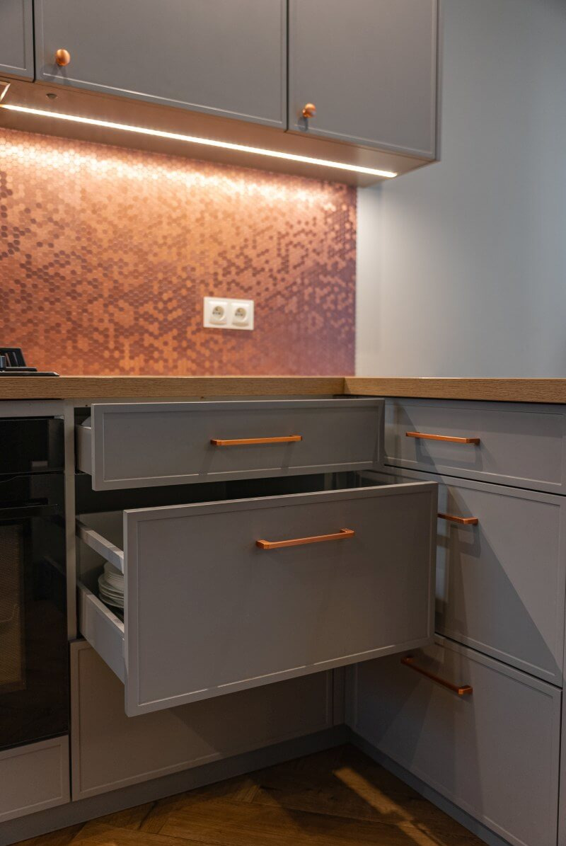 Szare szafki kuchenne z otwartymi szufladami i miedzianymi uchwytami, na tle mozaiki w odcieniach miedzi i oświetlenia LED. Widoczny fragment blatu i podłogi.