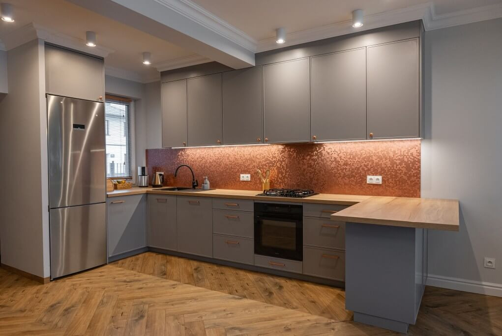 Szara kuchnia na wymiar z miedzianym panelem nad blatem i drewnianym blatem. Nowoczesne oświetlenie punktowe i parkiet w jodełkę. Lodówka ze stali nierdzewnej.
