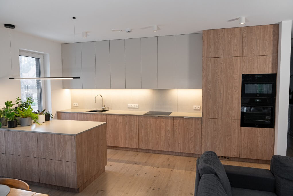 Nowoczesna kuchnia z wyspą. Jasne drewno i szare szafki górne. Oświetlenie LED pod szafkami. Minimalistyczny design z czarnymi akcentami. Widok na salon.