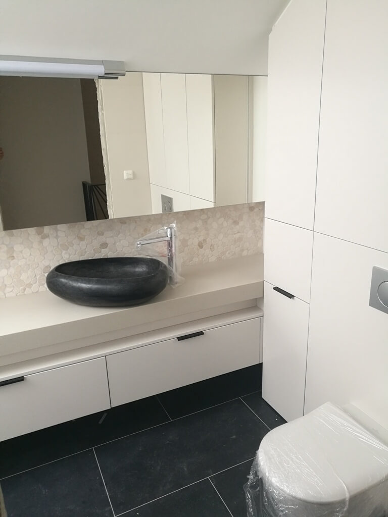 Nowoczesna łazienka z białą zabudową meblową, czarną umywalką i lustrem. Minimalistyczny design, czarne płytki podłogowe, toaleta w folii ochronnej.