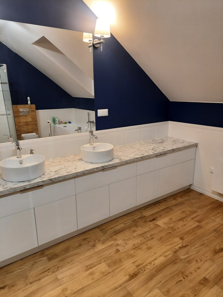 Białe szafki łazienkowe pod umywalki nablatowe z blatem imitującym marmur, na tle granatowej ściany i drewnianej podłogi. Ujęcie z perspektywy podłogi.