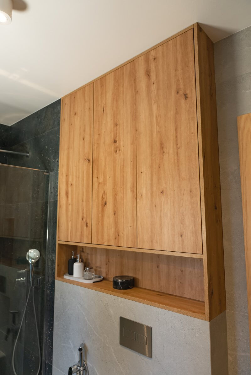 Drewniana szafka łazienkowa z otwartą półką, na której stoją akcesoria. Szafka w odcieniach brązu, na tle szarej ściany i kabiny prysznicowej.