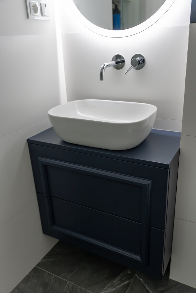 Granatowa szafka łazienkowa z umywalką nablatową i chromowaną armaturą, oświetlona okrągłym lustrem. Minimalistyczne wnętrze z szarymi płytkami podłogowymi.