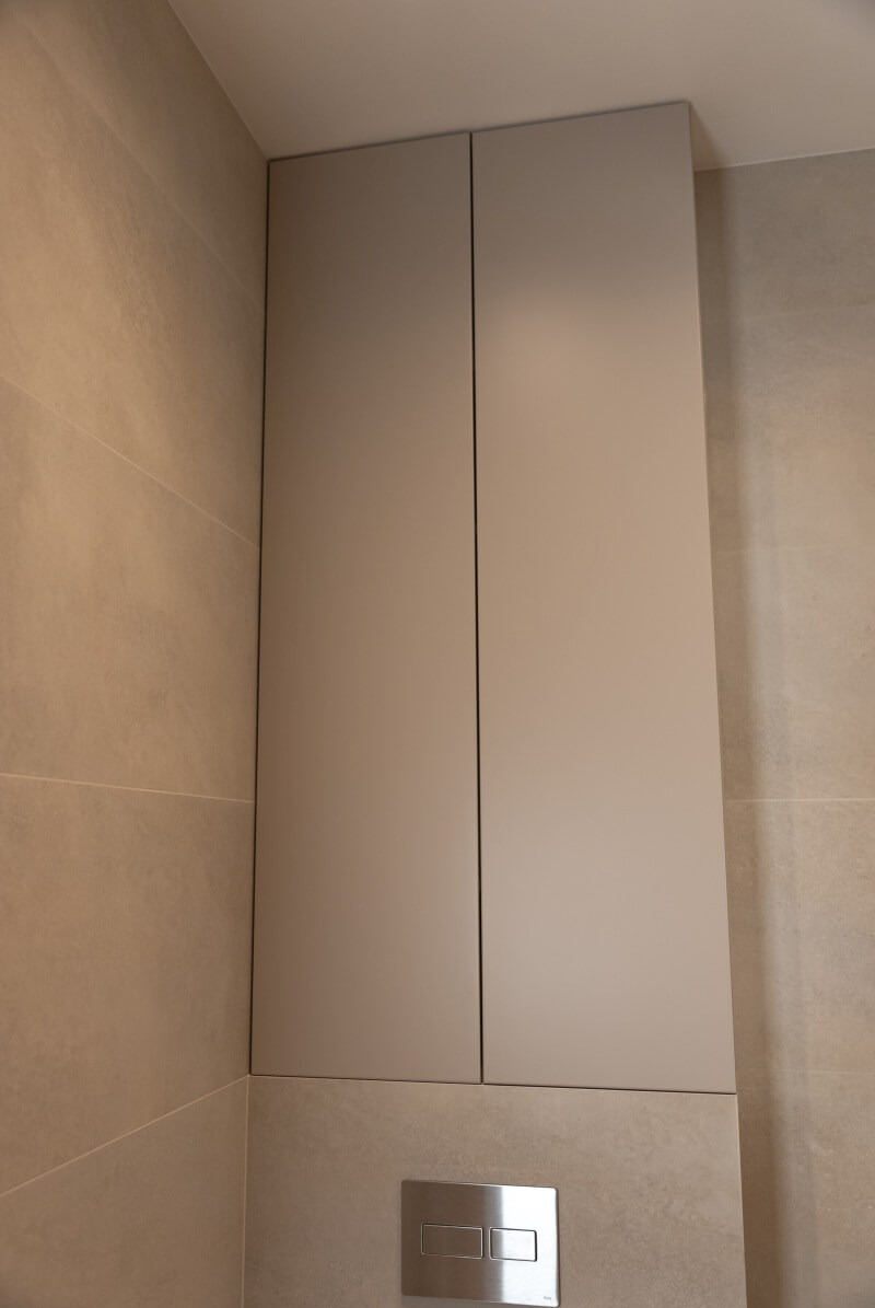 Beżowa szafka wnękowa nad toaletą z przyciskiem spłukującym, dopasowana kolorystycznie do płytek ściennych. Minimalistyczny design, proste linie.
